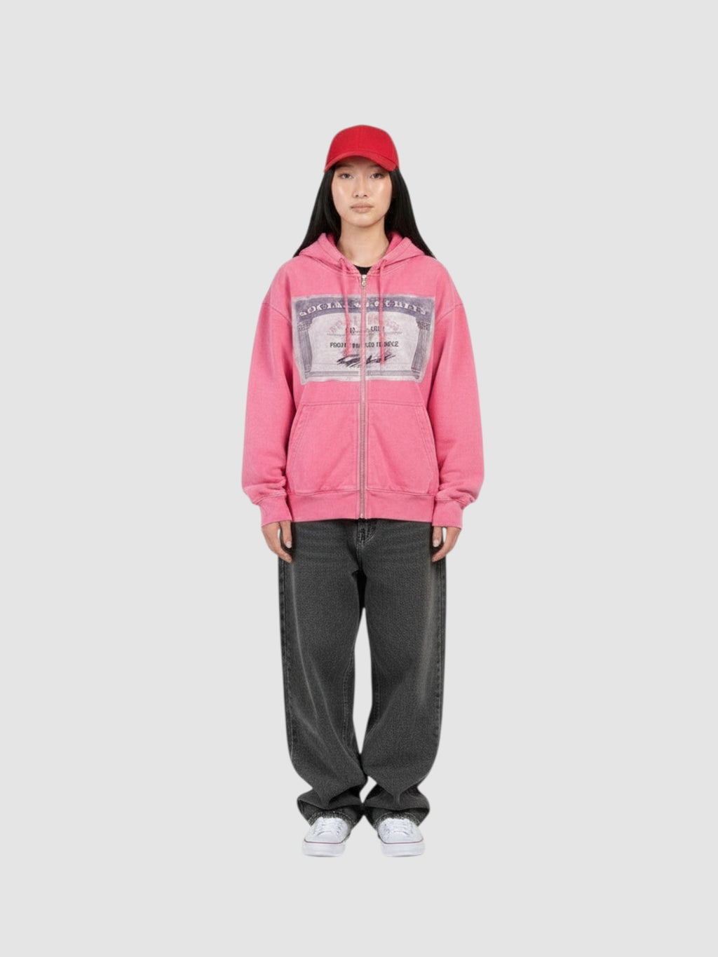 Y2K Studio pink zip-up hoodie with a graphic card print on the chest, worn by a model with a red cap and grey jeans.